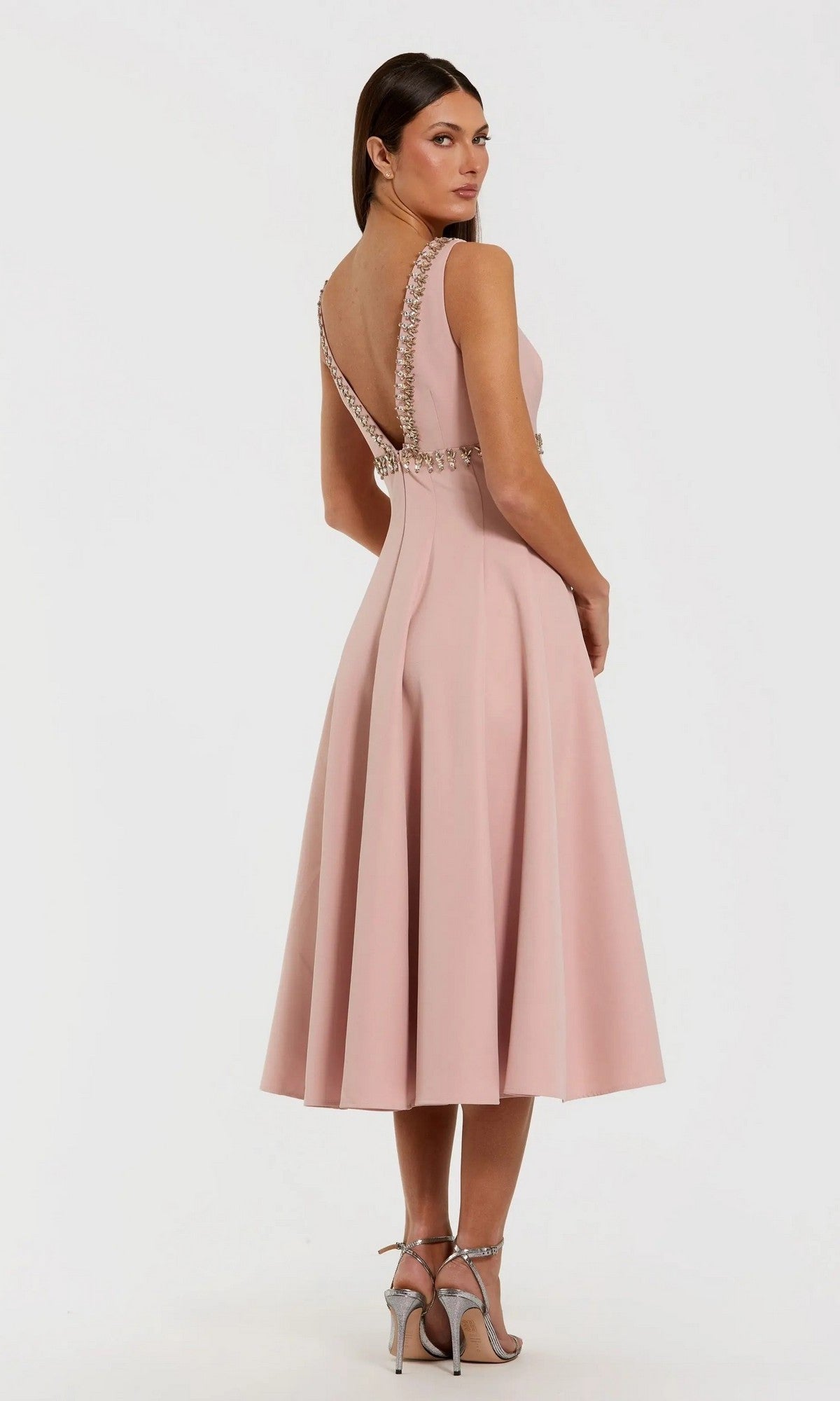 A woman stands against a white background wearing the Midi Wedding Guest Dress: Mac Duggal 21159, a sleeveless blush pink midi with a low V-back and decorative trim, paired elegantly with silver high-heeled sandals.